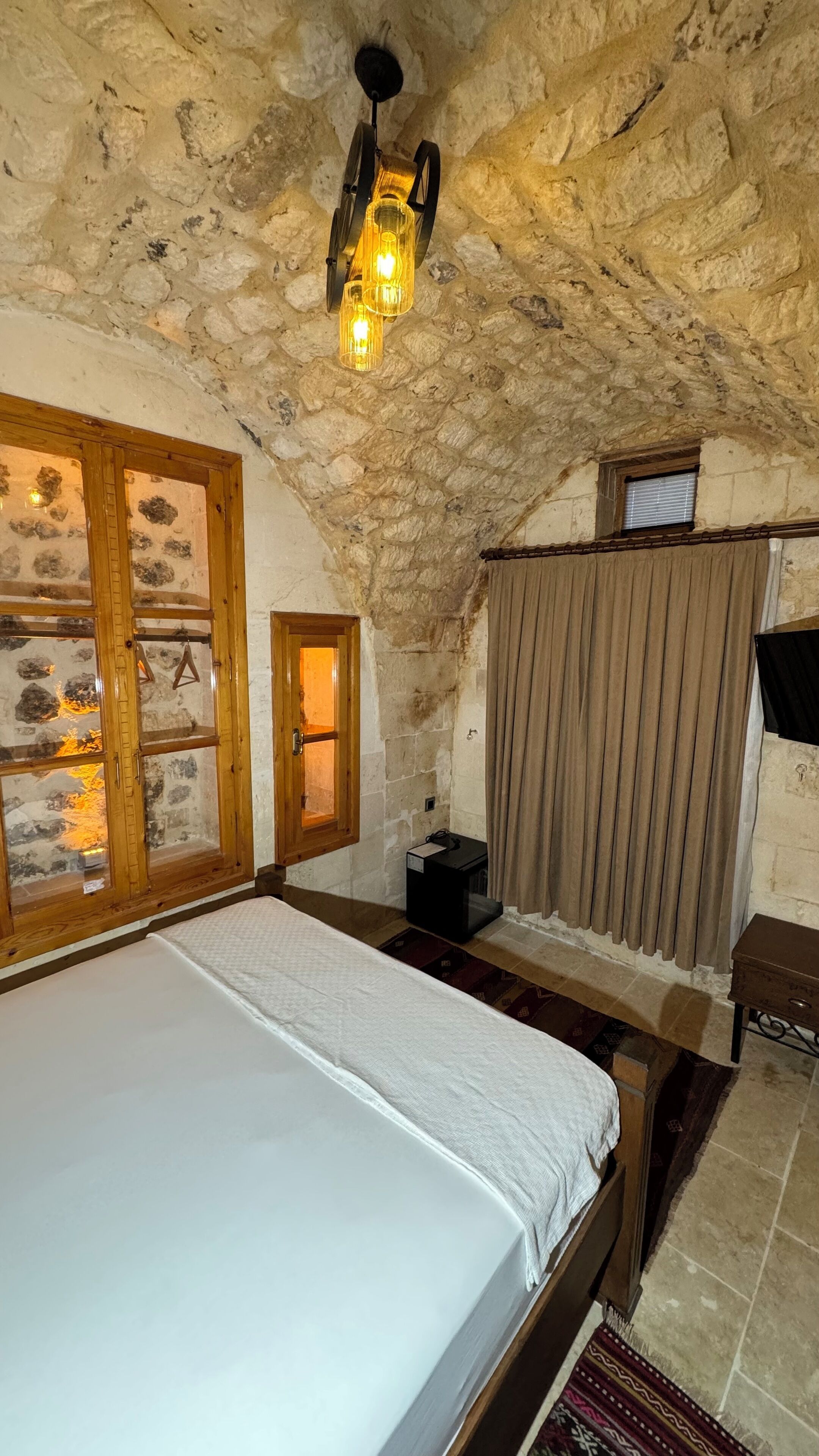 A rustic bedroom with a stone ceiling, a wooden bed, a stone fireplace, and a window with curtains.