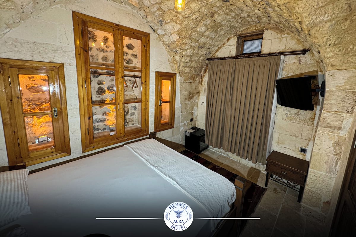 A stone-walled room with a bed, wooden-framed windows, a television, and a small table.