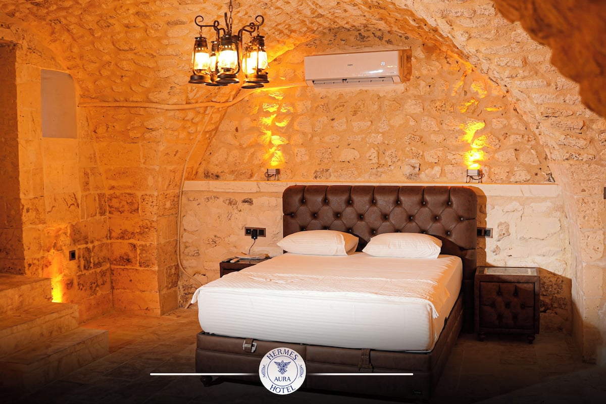 A bedroom with a stone wall, a bed, a nightstand, a lamp, and a ceiling light.
