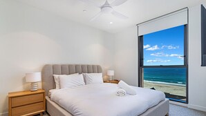Une chambre avec une grande fenêtre donnant sur la plage, un lit avec du linge de lit blanc, deux lampes de chevet et un ventilateur de plafond.