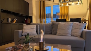 Un soggiorno moderno con un tavolino in vetro, un divano con cuscini, un caminetto e una zona cucina sullo sfondo.