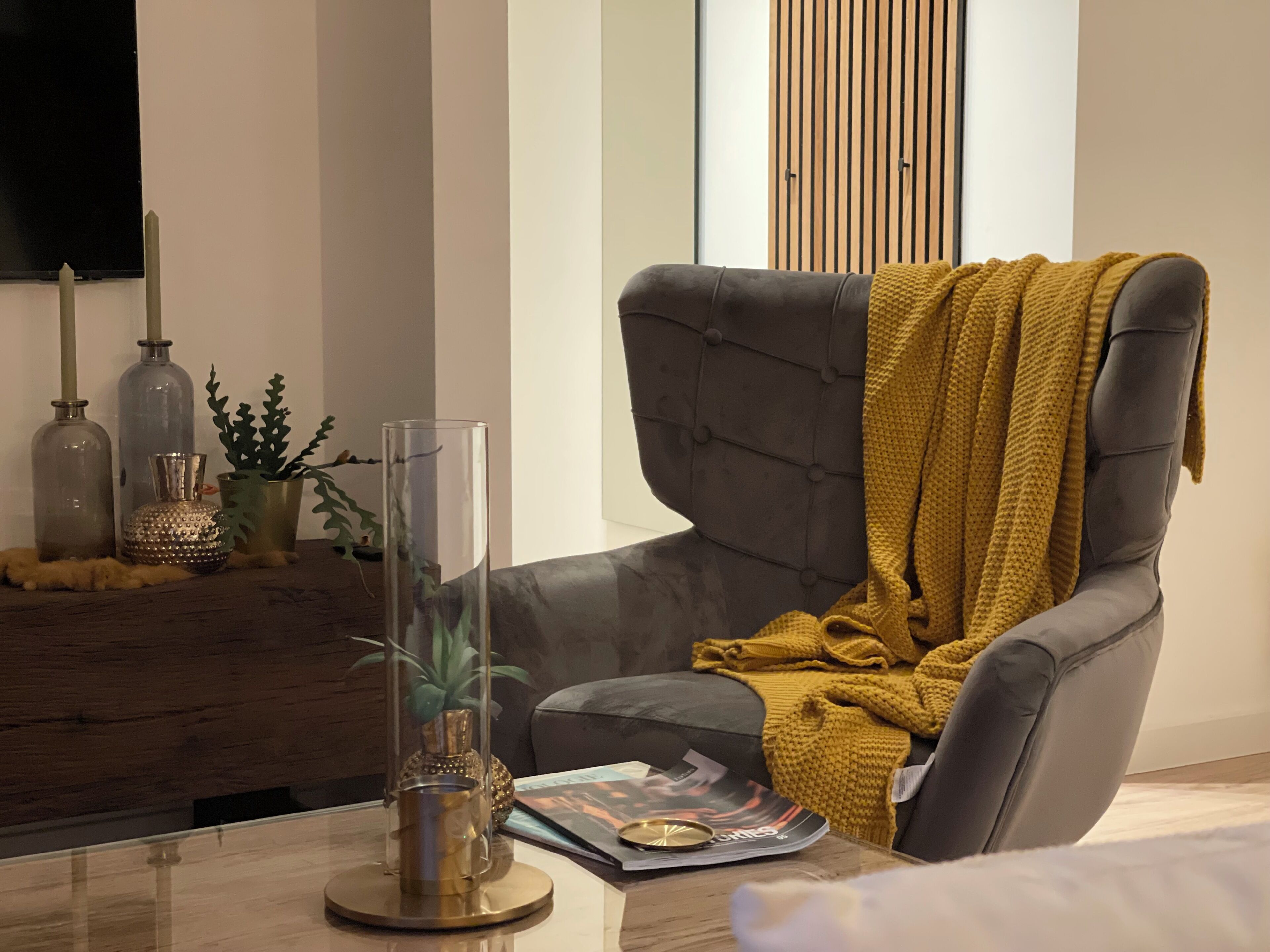 Een moderne woonkamer met een grijze fauteuil, een gele plaid, een glazen vaas met een plant en decoratieve flessen op een houten tafel.