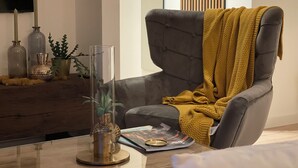 Un soggiorno moderno con una poltrona grigia, una coperta gialla, un vaso di vetro con una pianta e bottiglie decorative su un tavolino in legno.