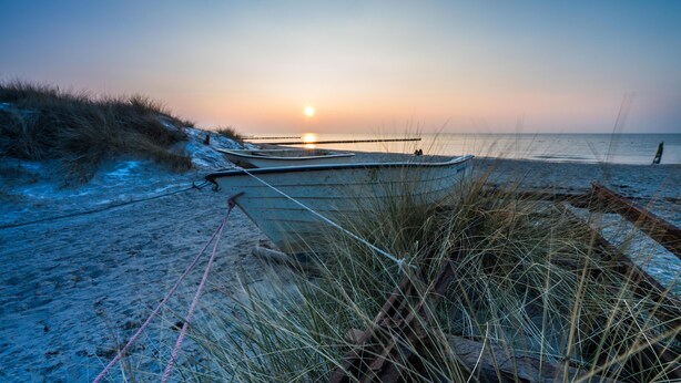 Una spiaggia con barche ormeggiate a un molo, un tramonto sullo sfondo e dune sabbiose.