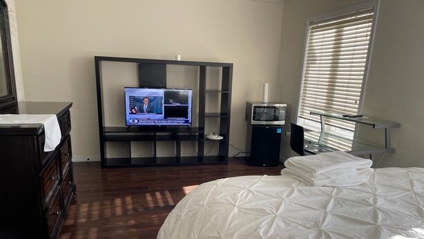 Ein Schlafzimmer mit Bett, Fernseher auf einem Schrank, Mikrowelle und einem Fenster mit Jalousien.