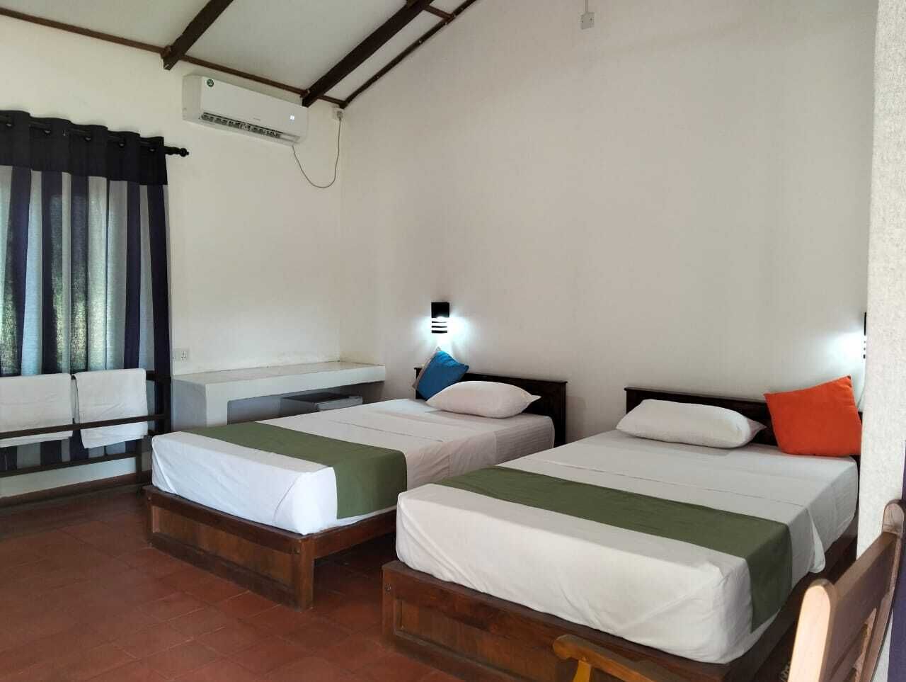 Une chambre avec deux lits simples, une petite table, un climatiseur fixé au mur et une fenêtre avec des rideaux.
