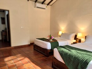 Camera d'albergo con due letti, pavimento in legno, un tavolino con un vaso e fiori, e condizionatore d'aria a muro.
