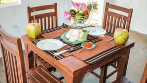 A wooden dining table with a placemat, two wooden chairs, a fruit basket, and a plate of food. - Tiara Elephant Lodge (Habarana)