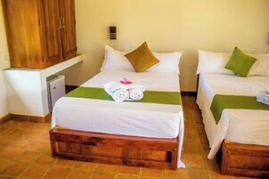 Habitación de hotel con dos camas, cada una con ropa de cama blanca y almohadones verdes. Hay un banco de madera al pie de las camas, y un pequeño escritorio con una lámpara.