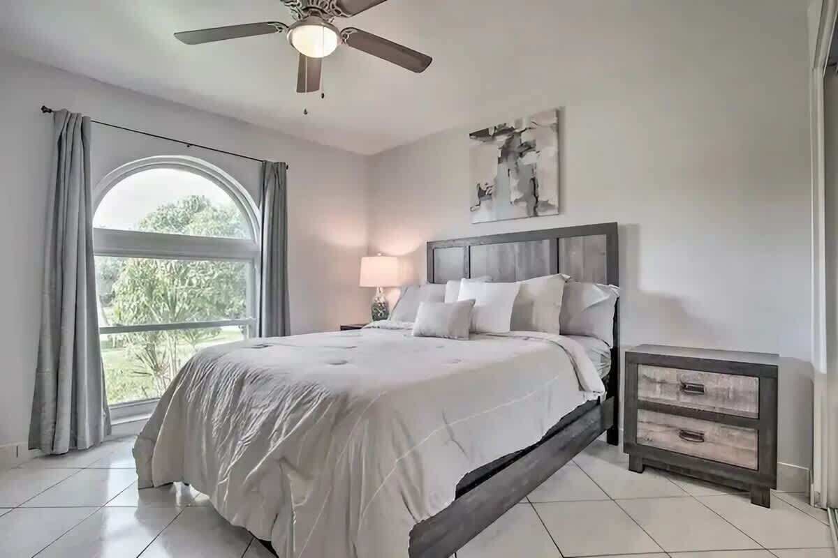 A bedroom with a bed, a nightstand, a ceiling fan, a window with curtains, and a wall-mounted artwork.