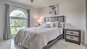A bedroom with a bed, a nightstand, a ceiling fan, a window with curtains, and a wall-mounted artwork.