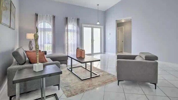 A modern living room with a grey sofa, a coffee table, and a lamp. There is a door leading to another room and a window with curtains.