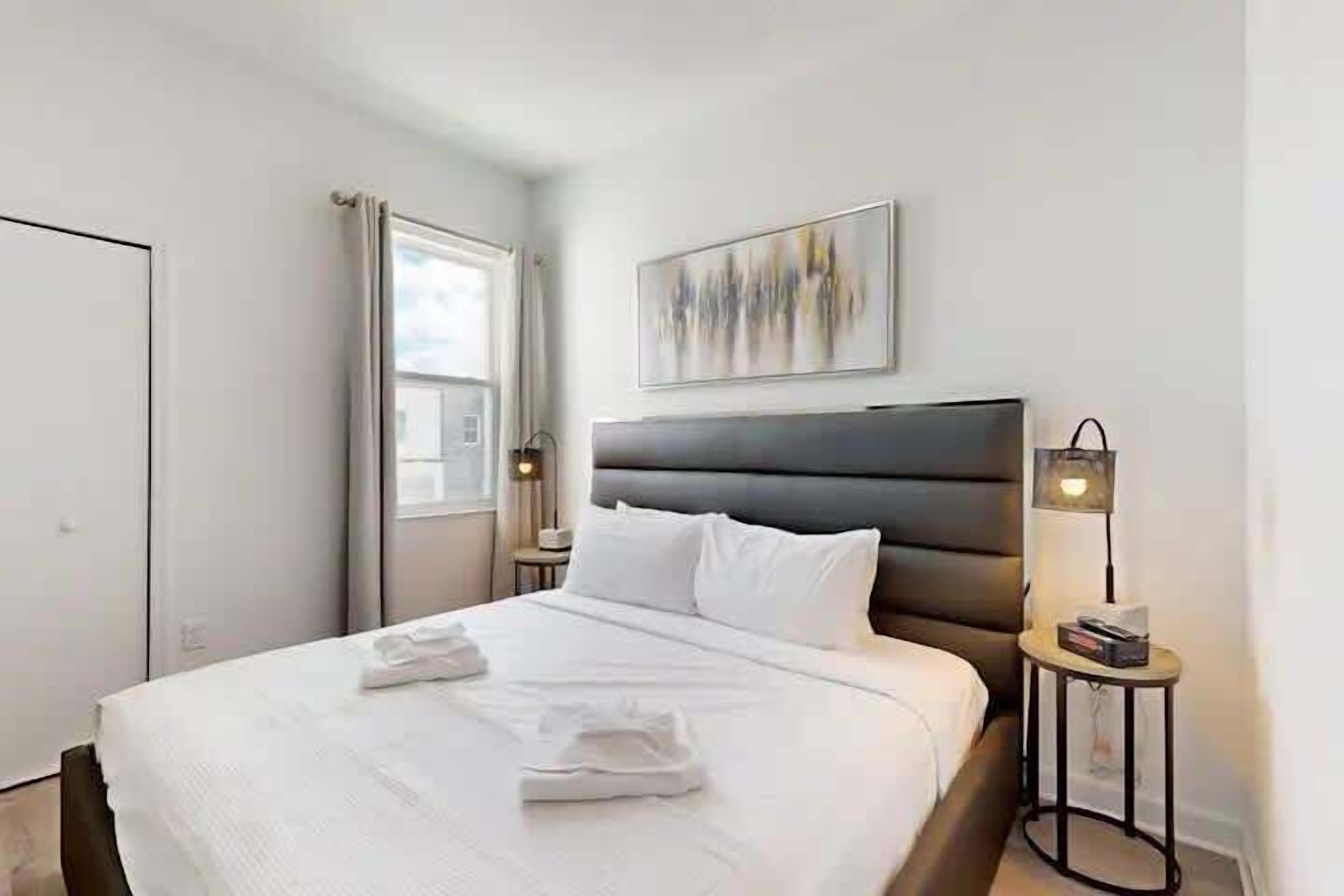 A modern hotel room with a large bed, a bedside table with a lamp, and a painting on the wall.