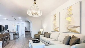 A modern living room with a white sofa, a coffee table, and a chandelier. There is a dining area with chairs and a table in the background. - 6BR Home w/ Private Pool, 15min to Disney + More! (Davenport)