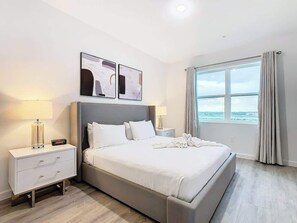 A modern hotel room with a large bed, bedside tables, and a view of the sea through a large window.