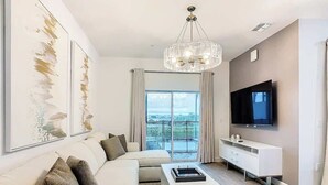 A modern living room with a white sofa, a coffee table, a TV, and a balcony with curtains. - 6BR Home w/ Private Pool, 15min to Disney + More! (Davenport)