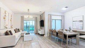 A modern living room with a white sofa, a coffee table, and a dining area with a table set for four. - 6BR Home w/ Private Pool, 15min to Disney + More! (Davenport)
