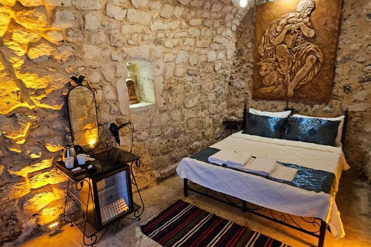A stone-walled room with a bed, a small table with a kettle, and a wall-mounted shelf.