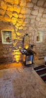 Habitación con paredes de piedra, una chimenea, una estantería metálica con tazas y dos ventanas pequeñas decoradas.