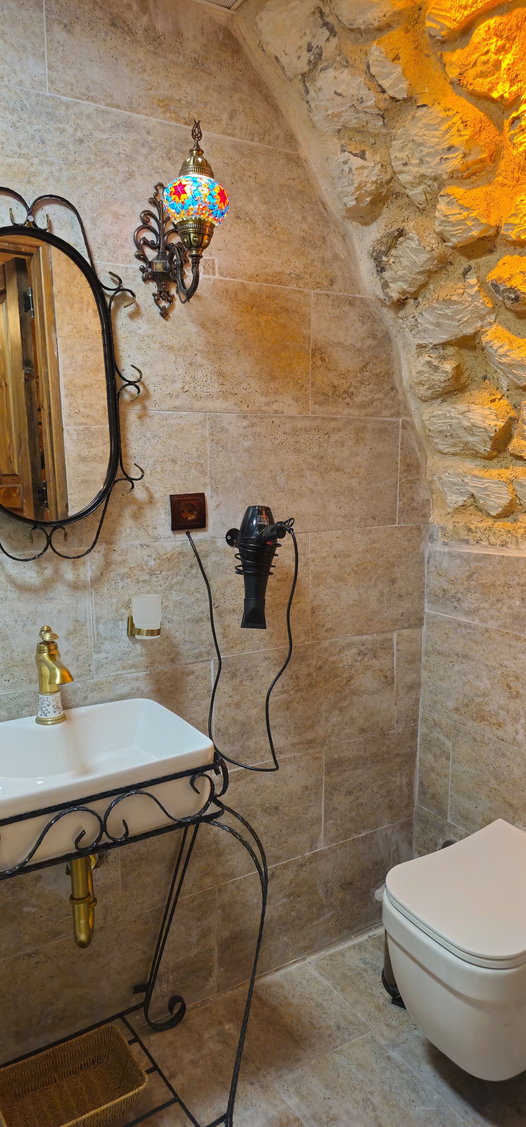 Un baño con pared de piedra, un lavamanos blanco con grifo dorado, un espejo, una lámpara decorativa y un inodoro.