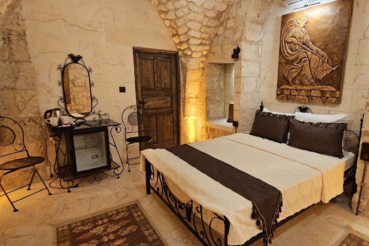A bedroom with a stone wall, a bed with dark bedding, a wooden door, a mirror, and a fireplace.