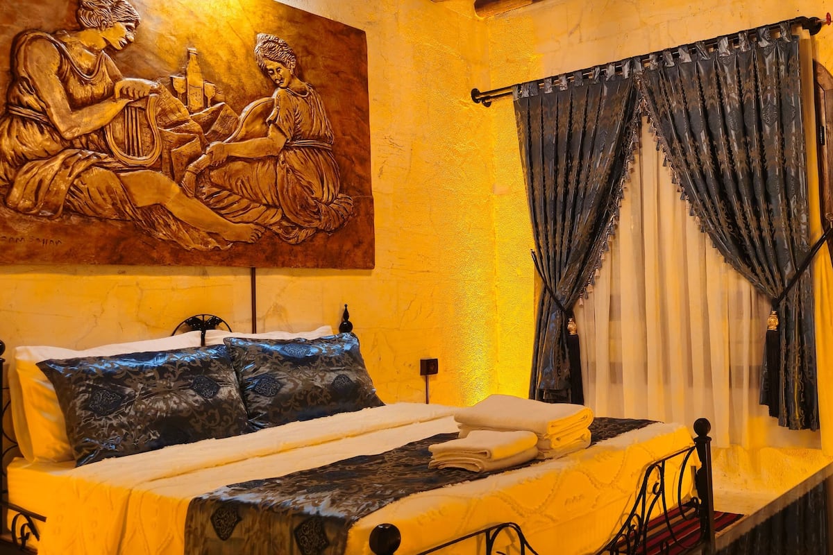 A bed with a headboard, a painting above it, a window with curtains, and a wooden door.