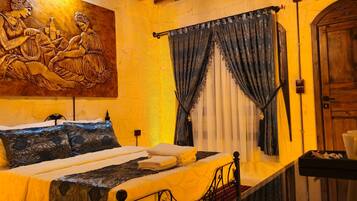 A bed with a headboard, a painting above it, a window with curtains, and a wooden door.