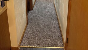 En smal hall med mattbeklädd golv och trädörrar, som leder till ett sovrum med en säng och ett nattduksbord.
