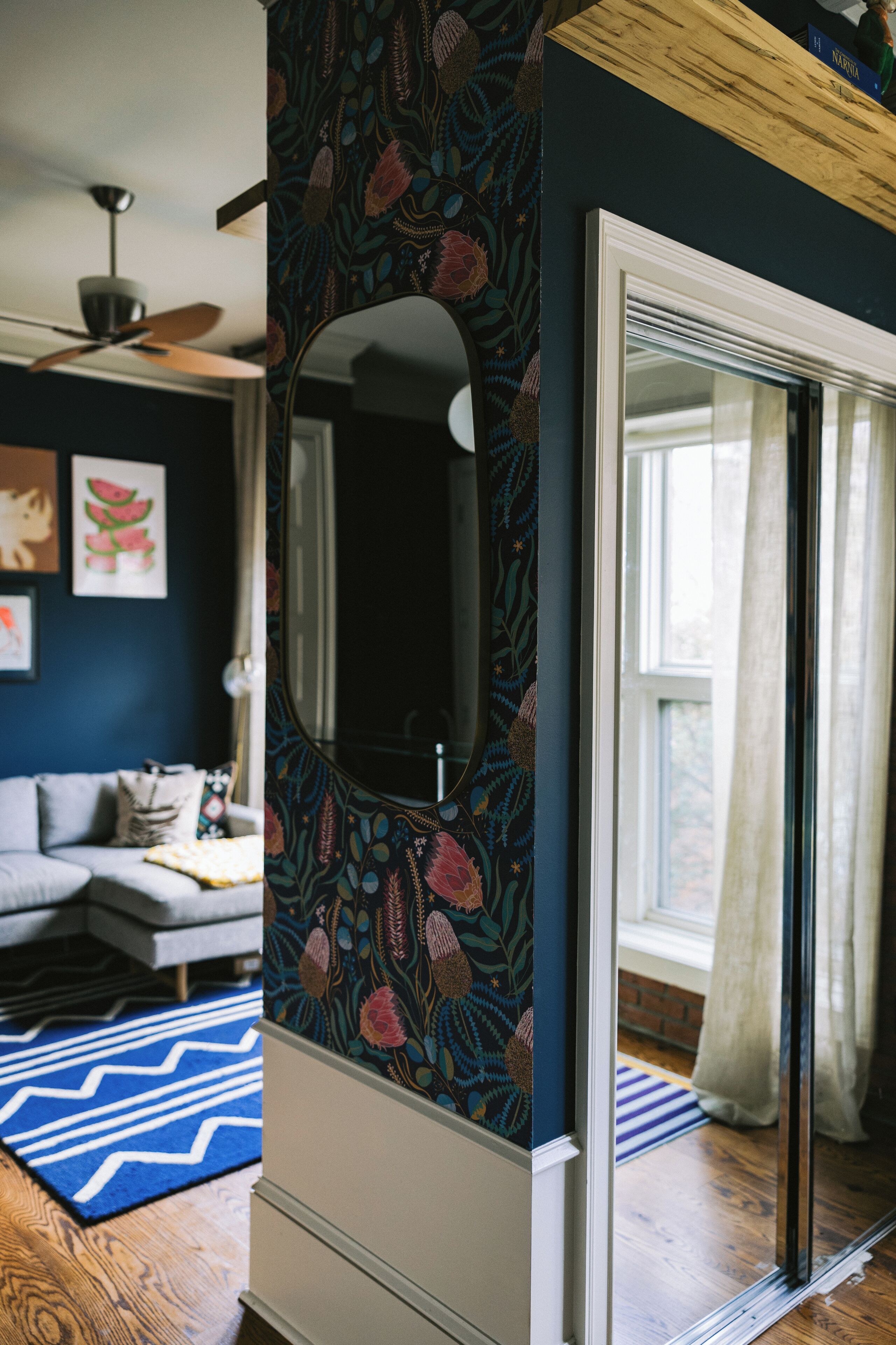 Une pièce avec un mur bleu foncé, un miroir, un ventilateur de plafond, un canapé et une fenêtre avec des rideaux.