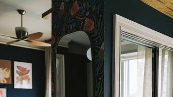 Une pièce avec un mur bleu foncé, un miroir, un ventilateur de plafond, un canapé et une fenêtre avec des rideaux.