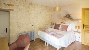 Un dormitorio con pared de piedra, cama con ropa blanca, una silla rosa y un radiador.