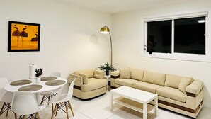 En stue med en beige sofa, et hvidt sofabord, et spisebord med hvide stole og en gulvlampe.
