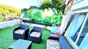 Un área de estar al aire libre con mobiliario de mimbre y césped artificial, con vistas a un jardín ajardinado con plantas tropicales.