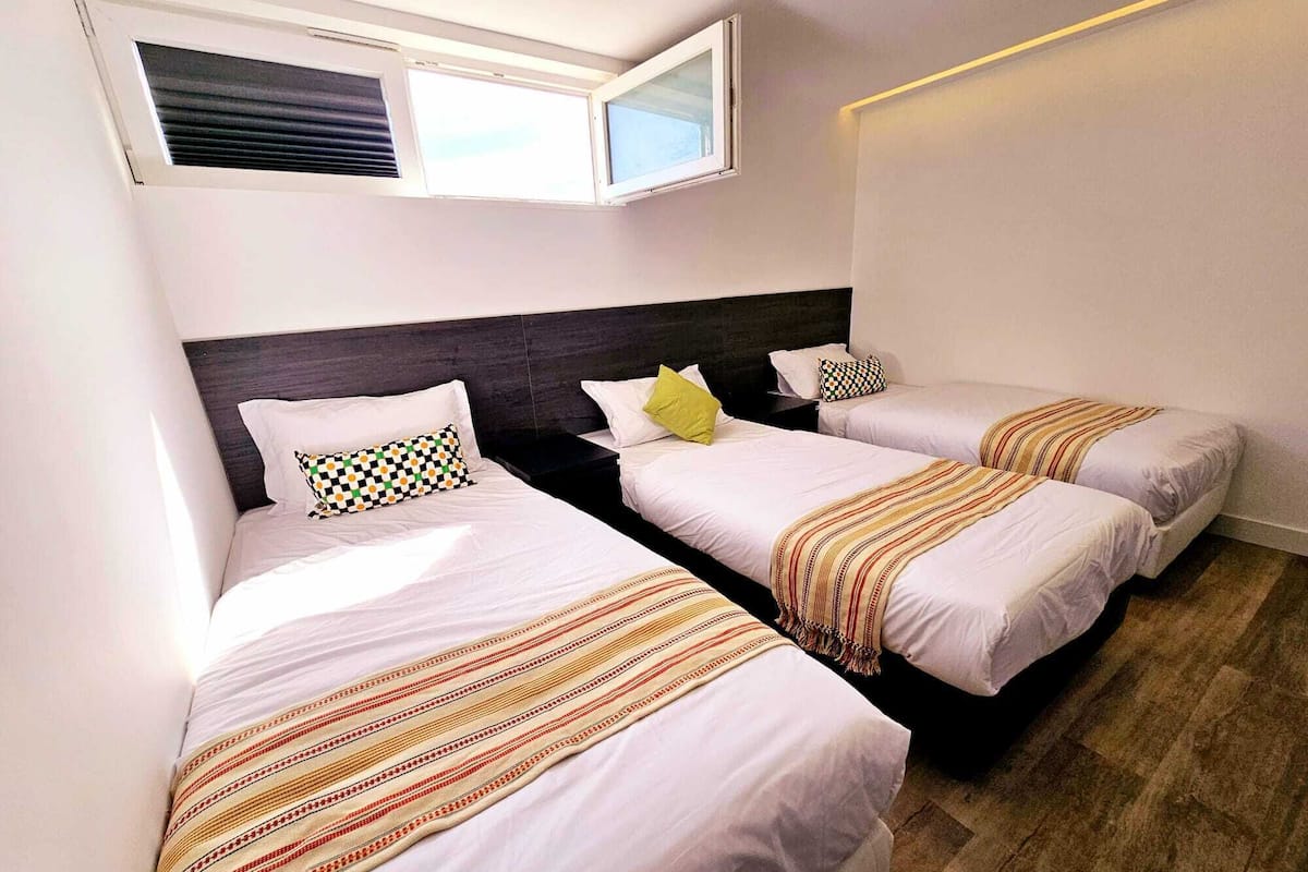 Ein Hotelzimmer mit drei Einzelbetten, jeweils mit einer gestreiften Decke und einem Kopfkissen. Oberhalb der Betten befindet sich ein Fenster, und der Boden ist mit Holz versehen.