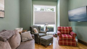 A living room with a grey sofa, a red plaid armchair, a grey armchair, a small table with a lamp, and a TV mounted on the wall.