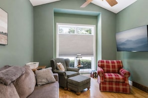Una sala de estar con un sofá gris, un sillón rojo a cuadros, otro sillón gris, una mesita con una lámpara y un televisor montado en la pared.