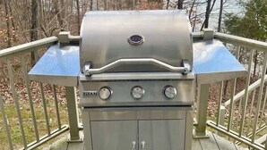 A stainless steel TITAN grill with three burners and a side shelf, located on a wooden deck with a railing, overlooking a forested area.