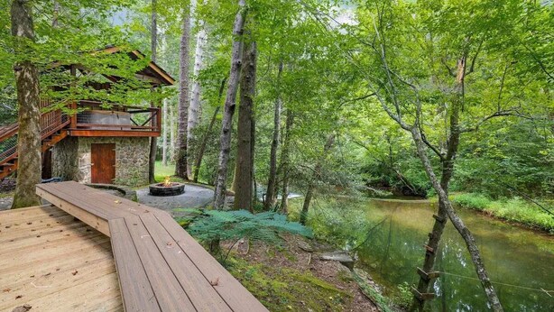 Una terraza de madera que lleva a una cabaña rústica con base de piedra, rodeada por un bosque frondoso y un río tranquilo.