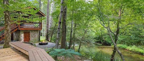 Una terraza de madera que lleva a una cabaña rústica con base de piedra, rodeada por un bosque frondoso y un río tranquilo.