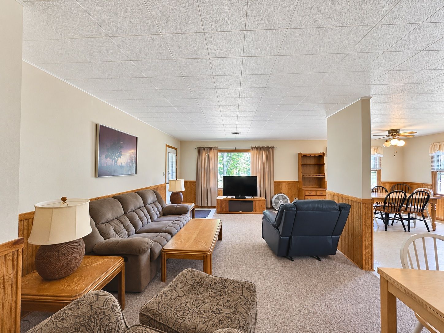 A spacious living room with a sofa, armchair, and a television. There is a dining area with a table and chairs.