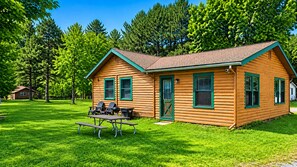 A wooden cabin with a picnic table and chairs, surrounded by a grassy area and trees.