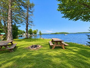 A lakeside picnic area with two wooden tables and a fire pit.