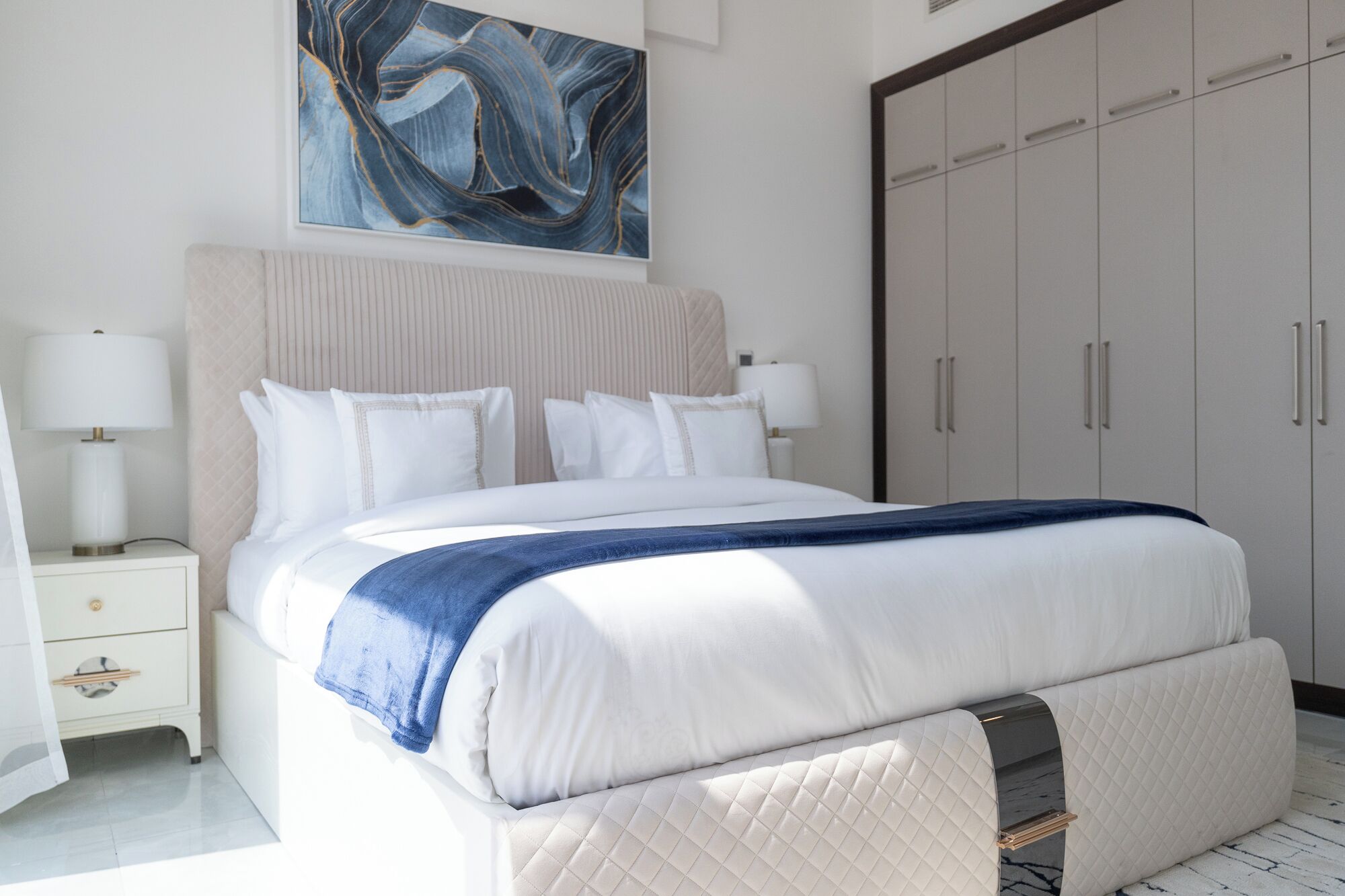 A modern bedroom with a large bed, white bedding, a blue throw blanket, and a headboard. There is a wardrobe with multiple doors and a painting above the bed.