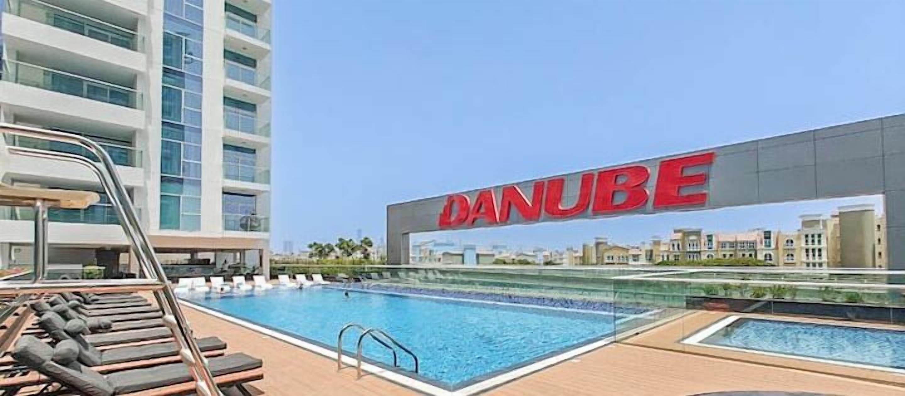 Een modern hotel met een groot buitenzwembad, een heldere lucht en de naam 'DANUBE' duidelijk zichtbaar.