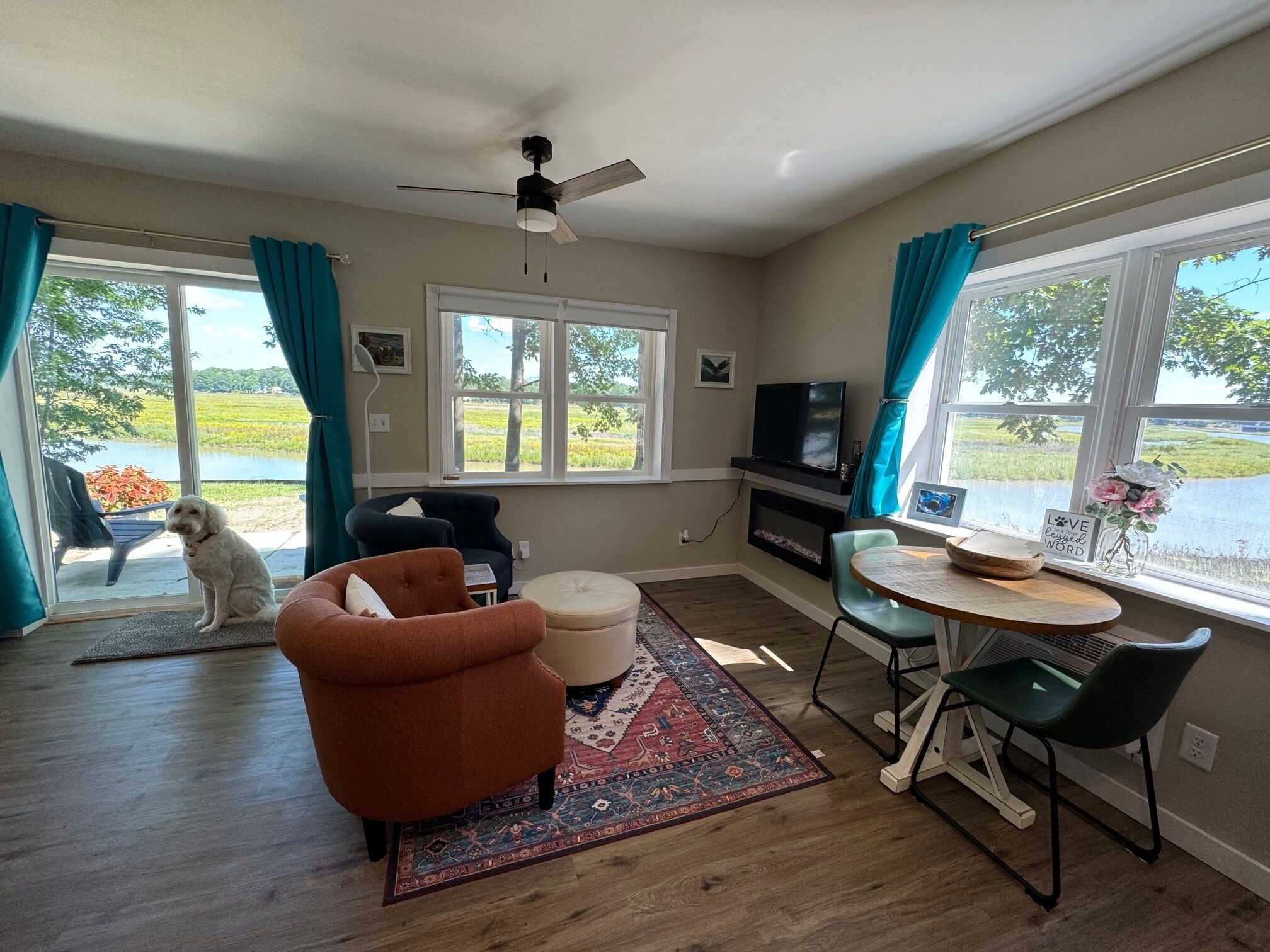 A living room with a sofa, armchair, ottoman, and a small table with chairs. There is a television and a fireplace. The room has large windows with curtains and a view of a lake.