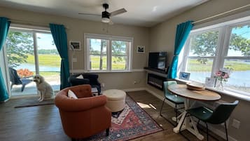 A living room with a sofa, armchair, ottoman, and a small table with chairs. There is a television and a fireplace. The room has large windows with curtains and a view of a lake.