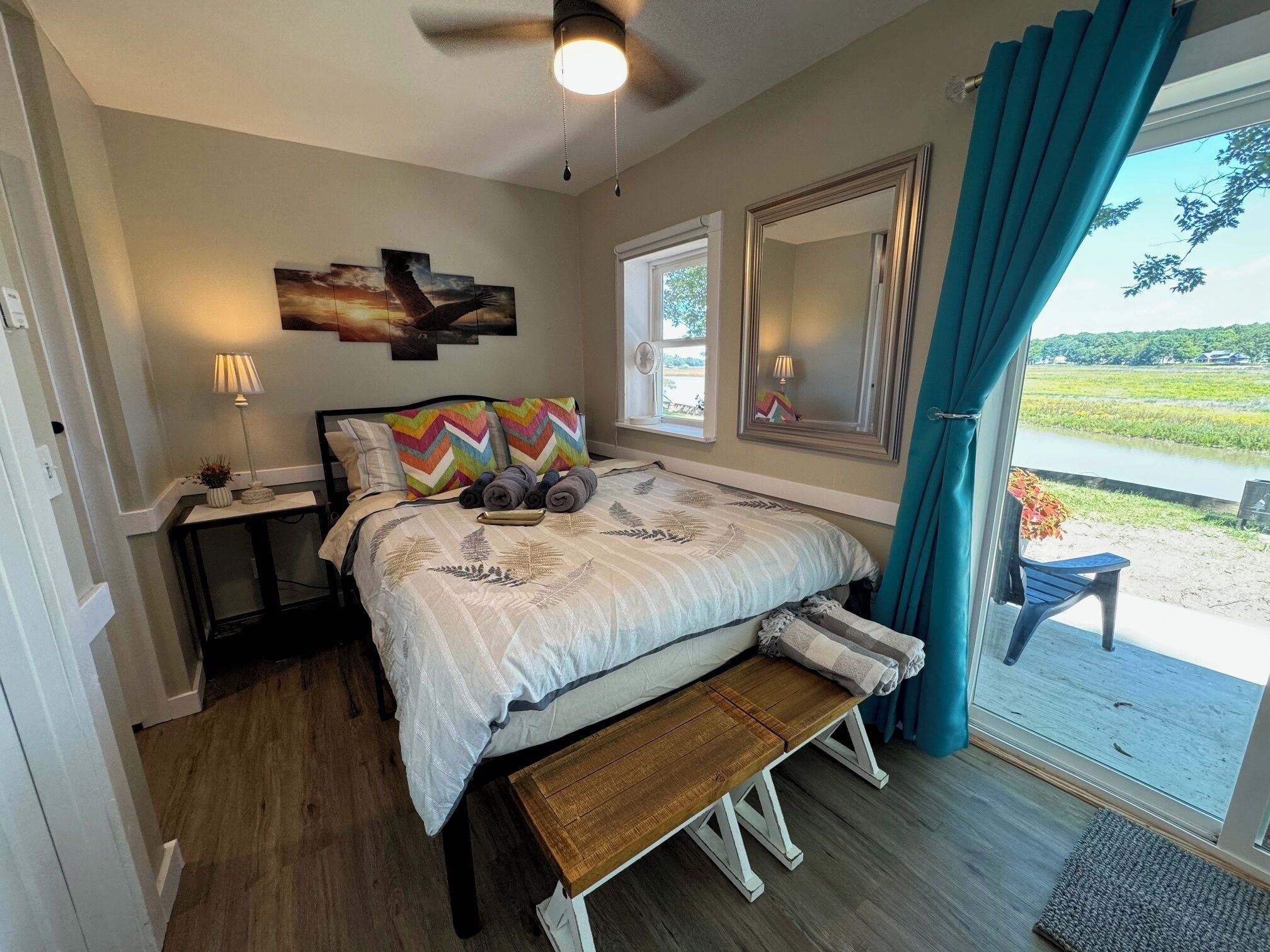Ein Schlafzimmer mit einem Bett, Nachttischen, einer Bank, einem Deckenventilator und Blick auf ein Gewässer und Grünflächen.