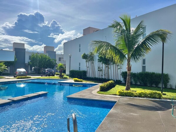 Una piscina con agua cristalina azul, rodeada por un jardín cuidado con palmeras y arbustos.