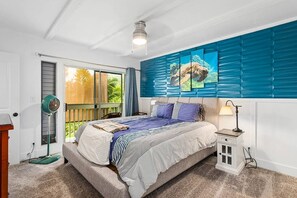 Ein Schlafzimmer mit Bett, Nachttisch, Standventilator und einem Fenster mit Blick auf Grünflächen.