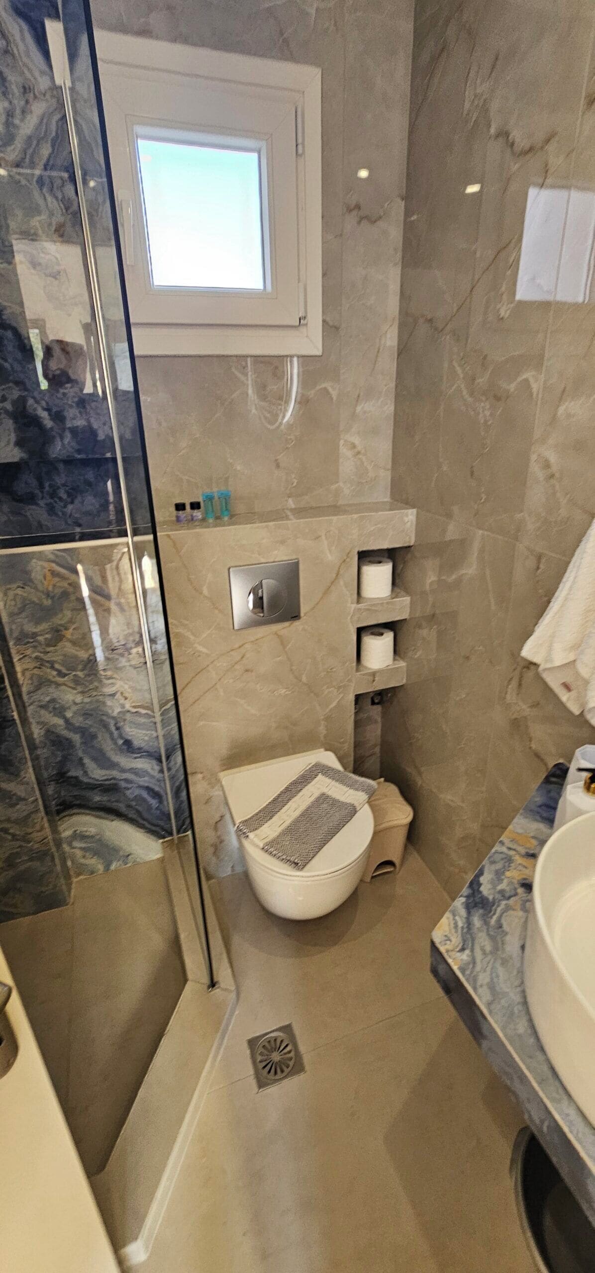 Een badkamer met een glazen douchecabine, toilet, wastafel en een wand betegeld met marmer.